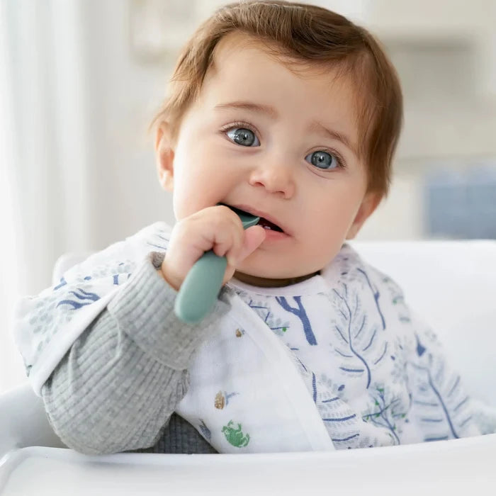 Baby holding a green teething ring and wearing a muslin bib with tree prints with a blurred background