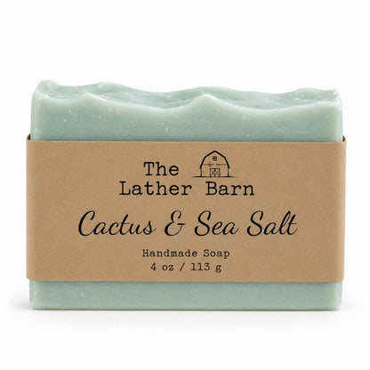 A handmade soap bar wrapped in a kraft paper band. The soap is a pale mint green with a wavy edge, and the label reads 'The Lather Barn Cactus & Sea Salt Handmade Soap 4 oz / 113 g'.