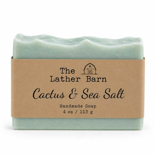 A handmade soap bar wrapped in a kraft paper band. The soap is a pale mint green with a wavy edge, and the label reads 'The Lather Barn Cactus & Sea Salt Handmade Soap 4 oz / 113 g'.