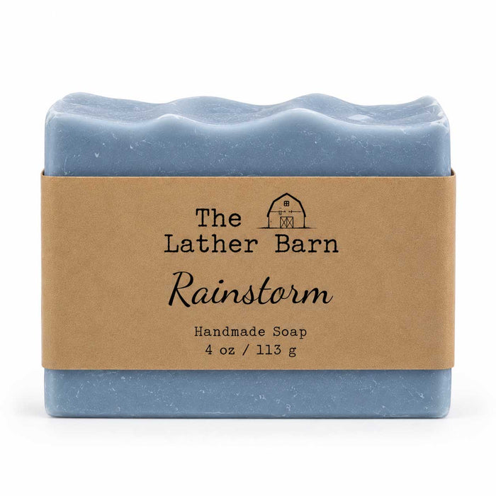 A rectangular bar of handmade soap in a dusty light blue color, wrapped in a brown kraft paper band with the brand name 'The Lather Barn' and product name 'Rainstorm' printed on it, set against a plain white background.
