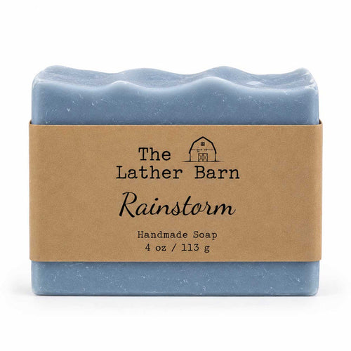 A rectangular bar of handmade soap in a dusty light blue color, wrapped in a brown kraft paper band with the brand name 'The Lather Barn' and product name 'Rainstorm' printed on it, set against a plain white background.
