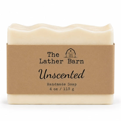 A rectangular bar of handmade soap in a cream-colored wrapper with a brown kraft paper label. The label reads 'The Lather Barn Unscented Handmade Soap 4 oz / 113 g' with a small barn logo.