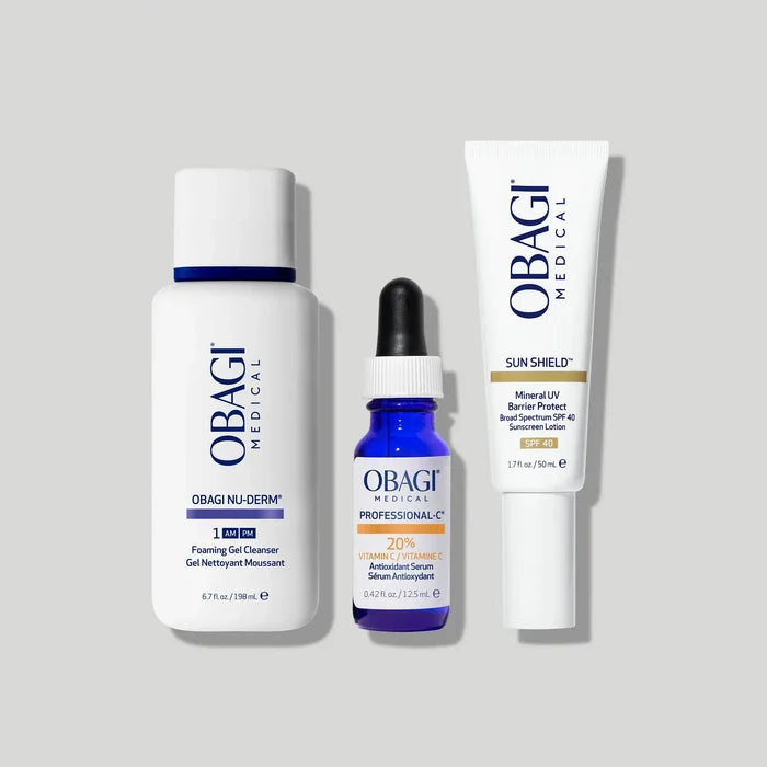 Three Obagi Medical skincare products arranged on a light gray background: a large white foaming gel cleanser bottle, a small blue vitamin serum dropper bottle, and a white sun shield tube.
